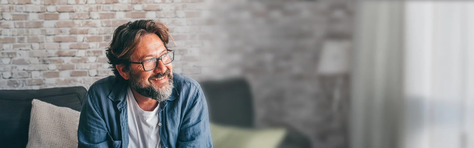 Mann sitzt im Wohnzimmer auf einem Sofa, schaut nachdenklich.