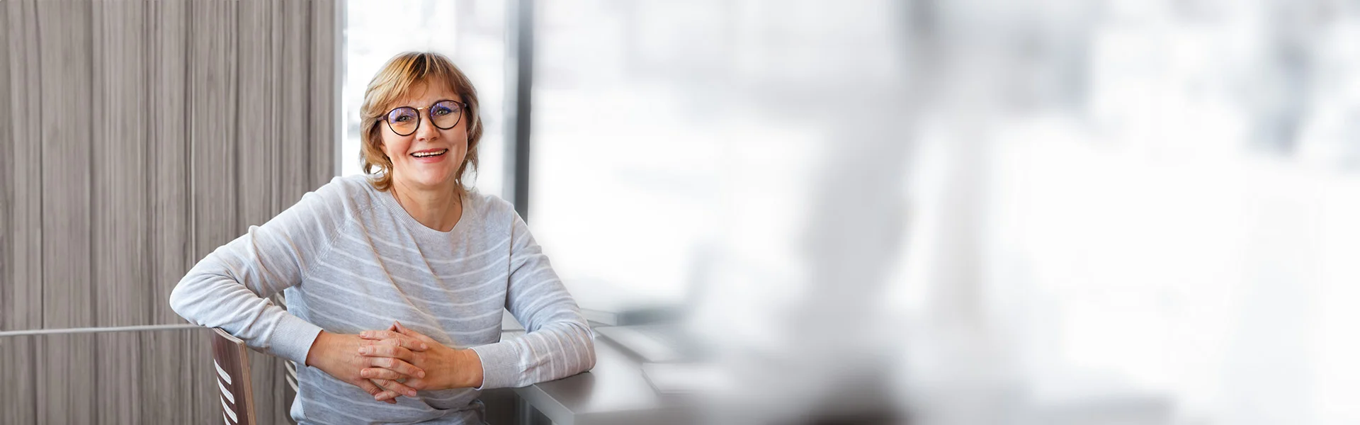 Frau mittleren Alters sitzt entspannt auf einem Stuhl und blickt freundlich in die Kamera.