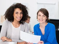 zwei Frauen sitzen an einem Tisch mit Papieren und lächeln in die Kamera