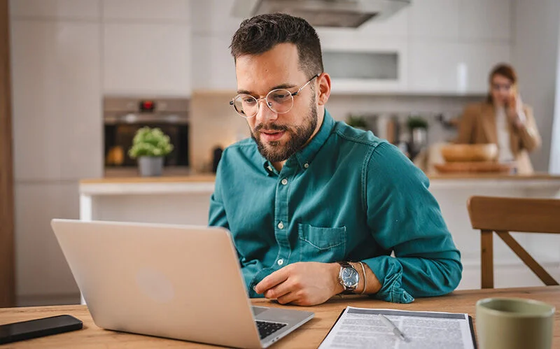 Mann lernt während seiner Weiterbildung im Homeoffice, während seine Frau in der Küche telefoniert.