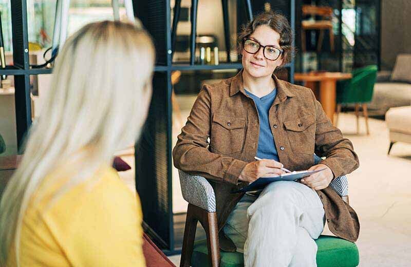 Blonde Frau schaut zu ihrer Coachin, die ihr aufmerksam zuhört und sich Notizen macht.