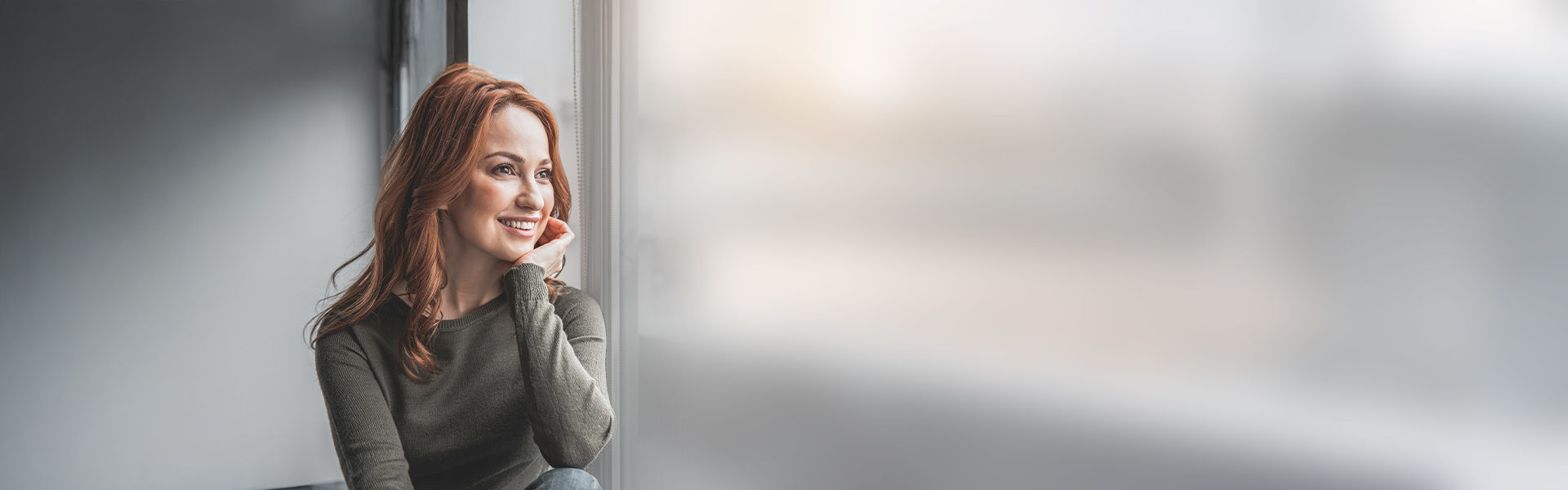 Junge Frau schaut lächelnd aus dem Fenster. Sie sieht freundlich aus.